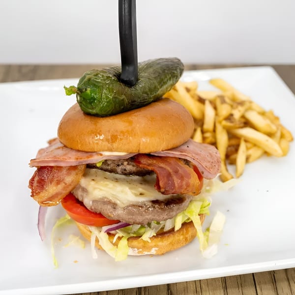 A large burger with bacon, ham, cheese, and vegetables, topped with a jalapeño and served with fries.