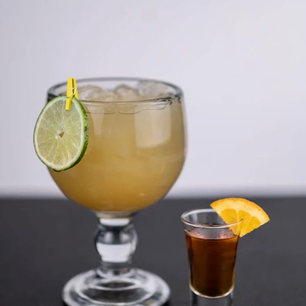 A large margarita with a lime slice and a shot of dark liquor with an orange slice.