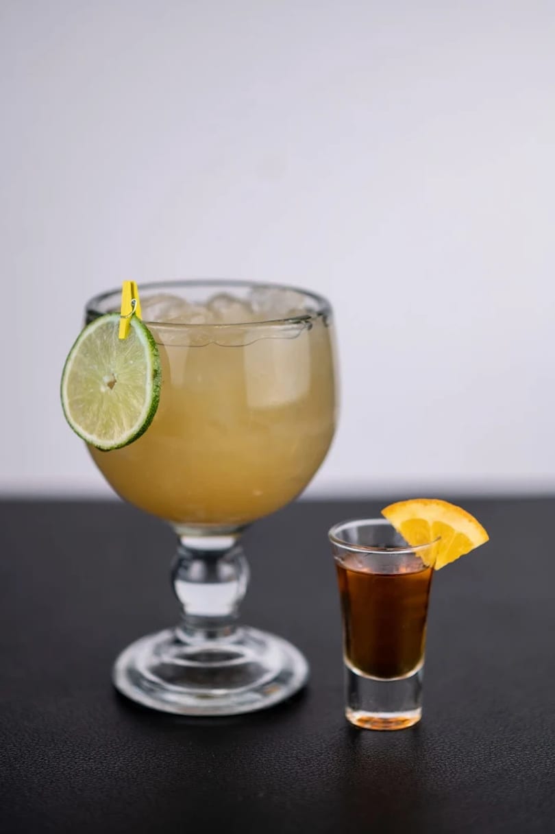 A large margarita with a lime slice and a shot of dark liquor with an orange slice.