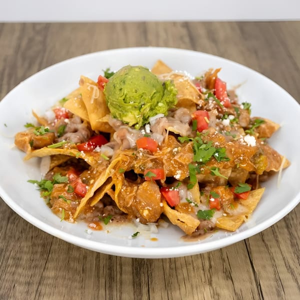 A white plate of nachos topped with guacamole, cheese, tomatoes, and cilantro on a wooden table.