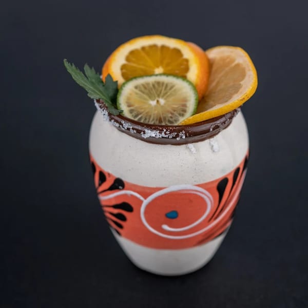 A colorful, handcrafted ceramic mug with a salted rim, garnished with orange, lemon, and lime slices.