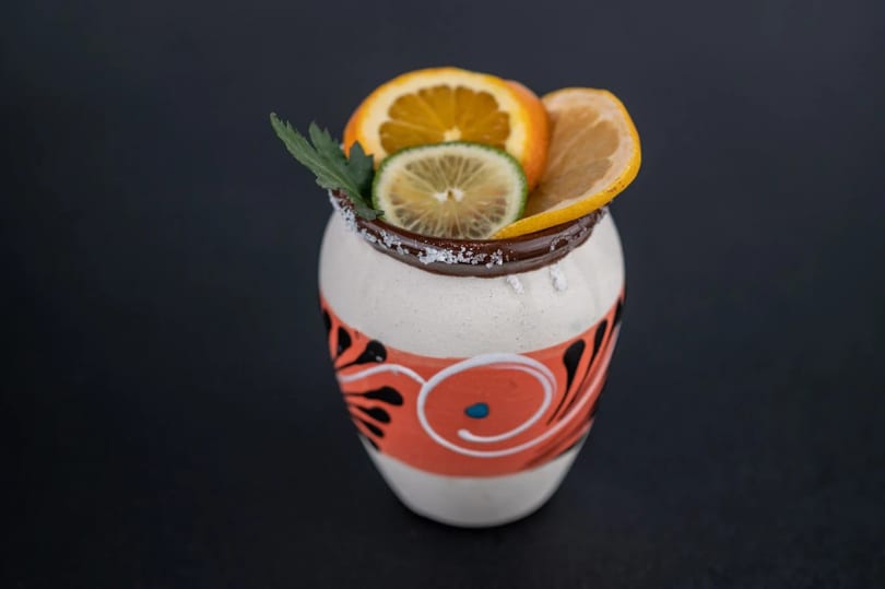 A colorful, handcrafted ceramic mug with a salted rim, garnished with orange, lemon, and lime slices.
