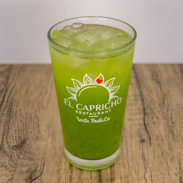 A tall glass of green iced beverage with the El Capricho Restaurant logo on a wooden table.