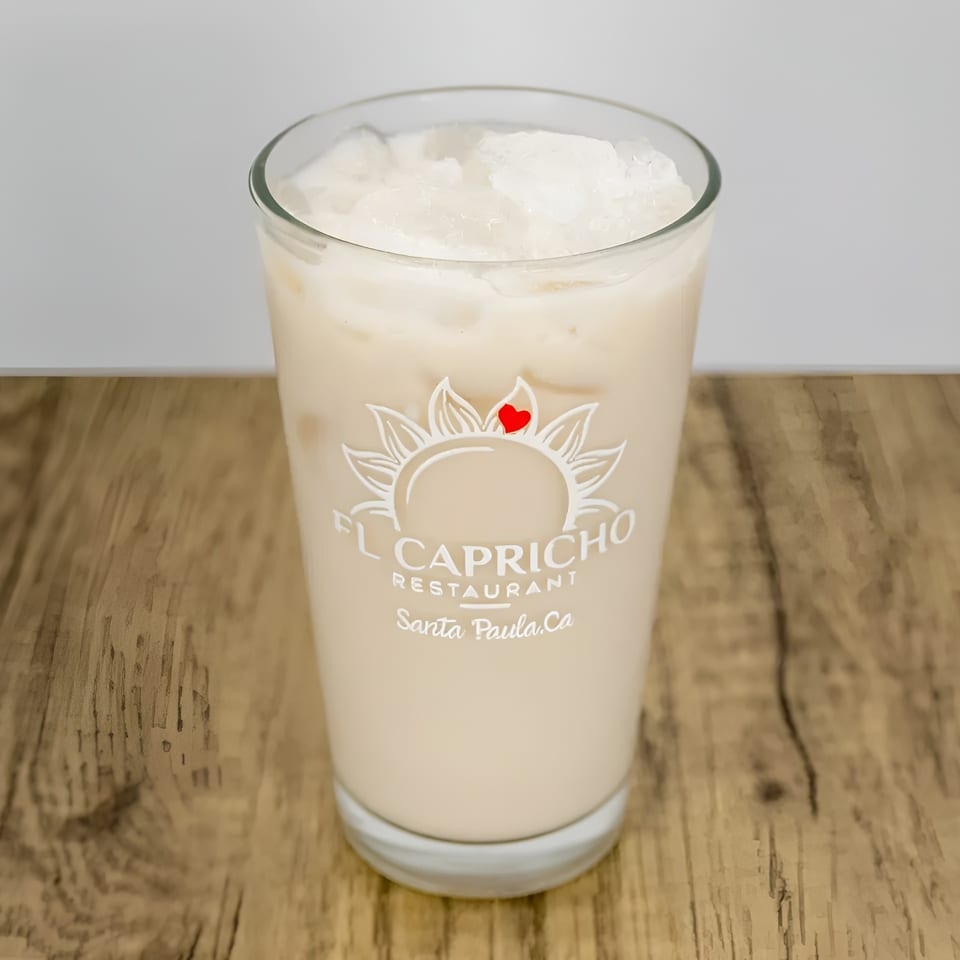 A glass of horchata with ice, featuring the El Capricho Restaurant logo, on a wooden table.