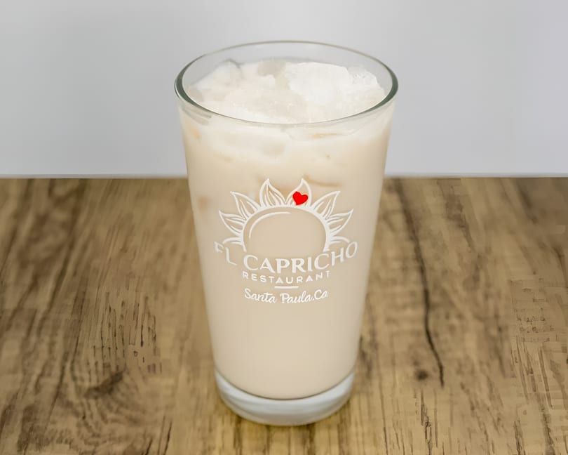 A glass of horchata with ice, featuring the El Capricho Restaurant logo, on a wooden table.