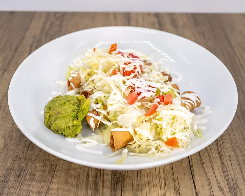 Taquitos topped with shredded lettuce, diced tomatoes, sour cream, and a side of guacamole.