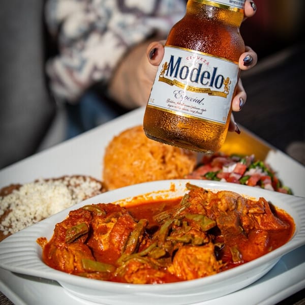 A hand holds a Modelo Especial beer bottle over a plate of Mexican food with red sauce.