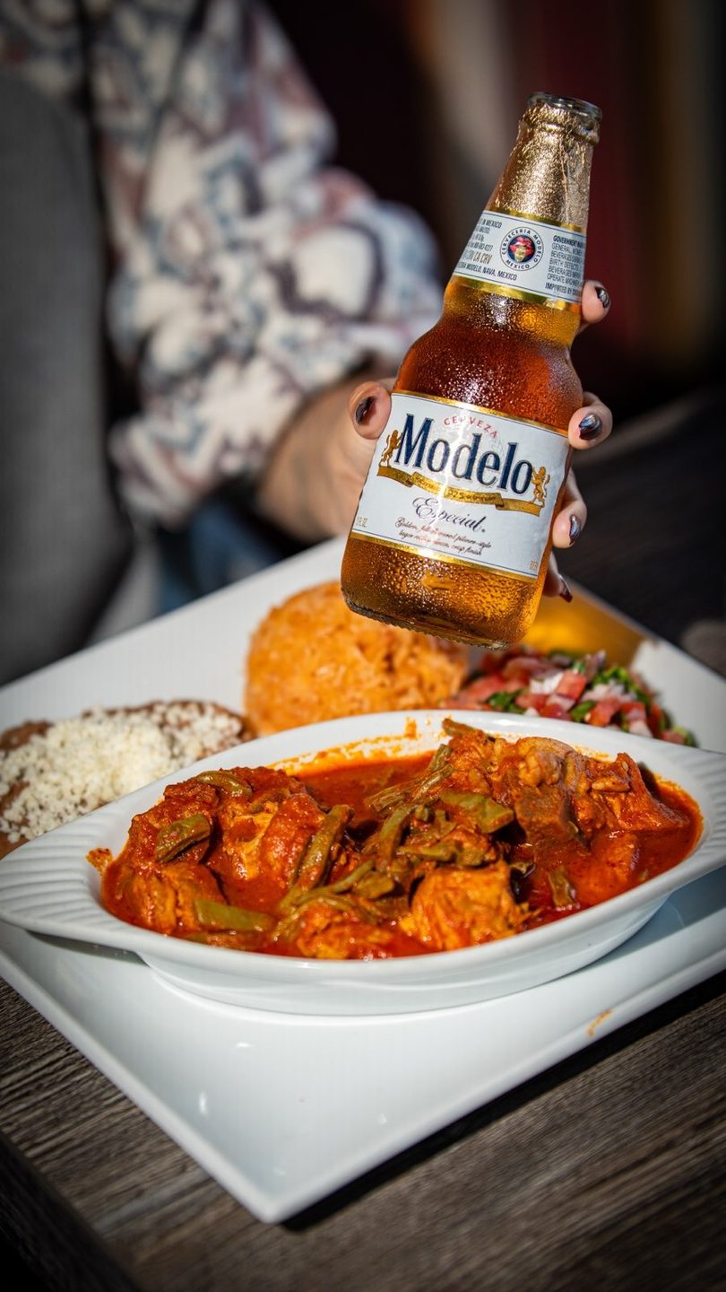 A hand holds a Modelo Especial beer bottle over a plate of Mexican food with red sauce.